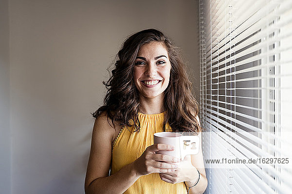 Beautiful woman holding coffee cup while standing by window blinds at home