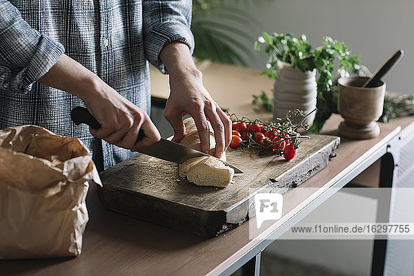 Hände eines Mannes,  der frisches,  hausgemachtes Brot auf einem Brett in der Küche schneidet