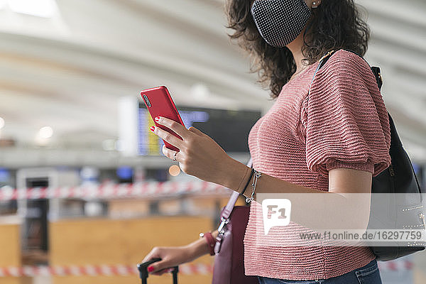 Nahaufnahme einer Frau mit Schutzmaske im Gesicht  die ein Smartphone benutzt  während sie am Flughafen steht