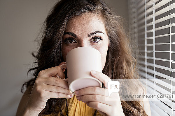 Nahaufnahme einer jungen Frau mit langen braunen Haaren  die zu Hause vor den Jalousien Kaffee trinkt