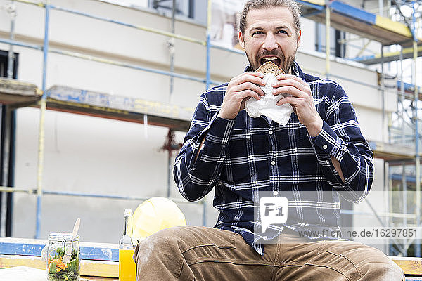Construction worker eating bread while sitting against building at construction site