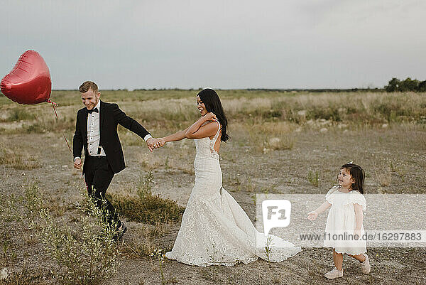Braut und Bräutigam mit herzförmigen Ballons halten sich an den Händen  während ihre Tochter im Feld spazieren geht