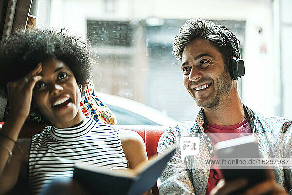 Smiling man listening music while girlfriend sitting with book against window in cafe