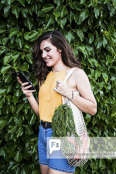 Glückliche junge Frau  die ihr Smartphone benutzt  während sie mit Gemüse in einem Netzbeutel vor grünen Pflanzen steht