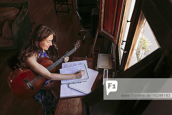 Junge Frau schreibt in ein Buch  während sie am Wohnzimmertisch Gitarre übt