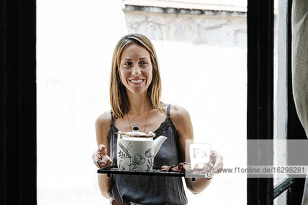 Smiling woman holding tray with kettle and dry flowers