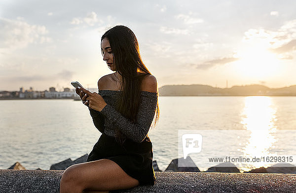 Junge Frau  die ein Mobiltelefon benutzt  während sie auf einer Stützmauer an der Promenade sitzt