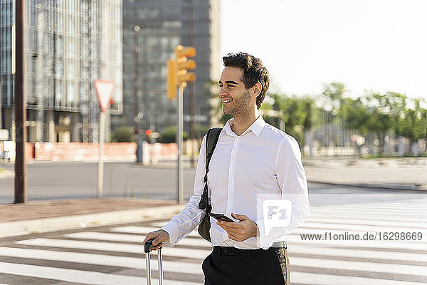 Lächelnder Geschäftsmann  der ein Smartphone und einen Koffer hält und wegschaut  während er auf einer Straße in der Stadt steht