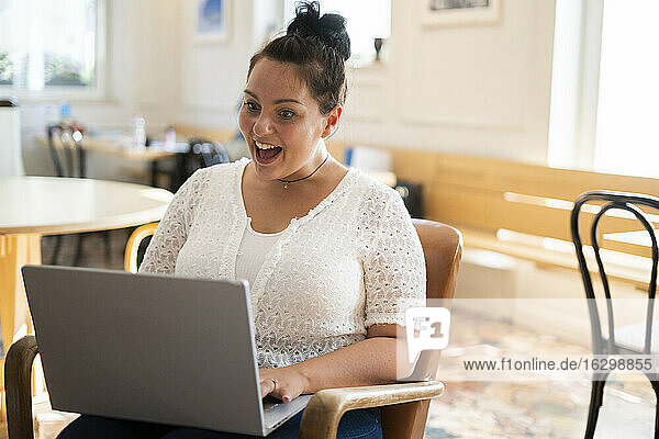 Schockierte junge Frau mit offenem Mund  die einen Laptop benutzt  während sie auf einem Stuhl in einem Café sitzt