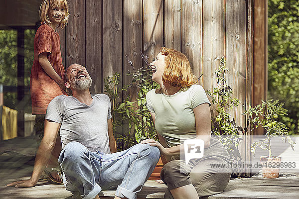 Mädchen neben fröhlichen Eltern  die lachend vor einem kleinen Haus sitzen