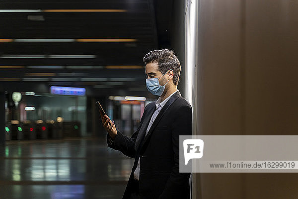 Männlicher Berufstätiger mit Maske  der ein Smartphone benutzt  während er an der Wand eines Bahnhofs steht