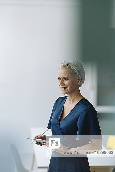 Lächelnde Unternehmerin mit digitalem Tablet  die wegschaut  während sie im Büro steht