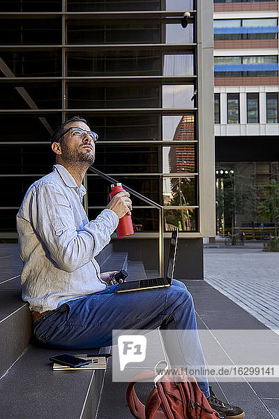 Businessman with laptop drinking water while looking up in city