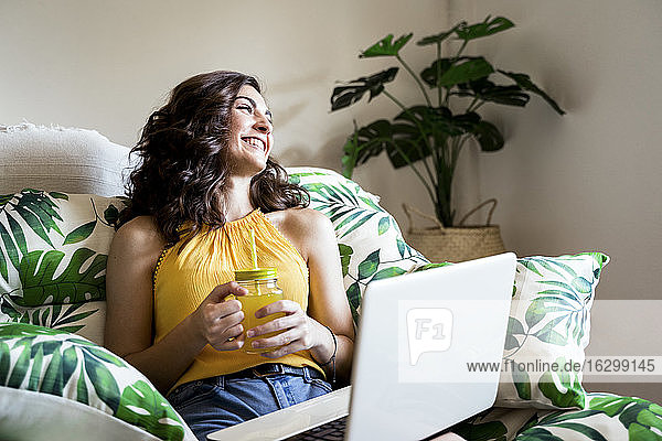 Cheerful young woman holding juice while sitting with laptop and looking away at home