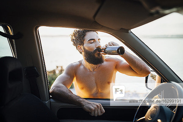 Shirtless bearded man drinking beer while leaning on car window