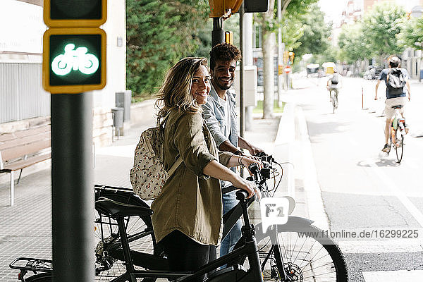 Smiling young couple with electric bicycles standing on sidewalk