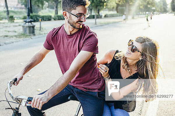 Fröhliche junge Frau sitzt mit ihrem Freund auf dem Fahrrad auf der Straße im Park