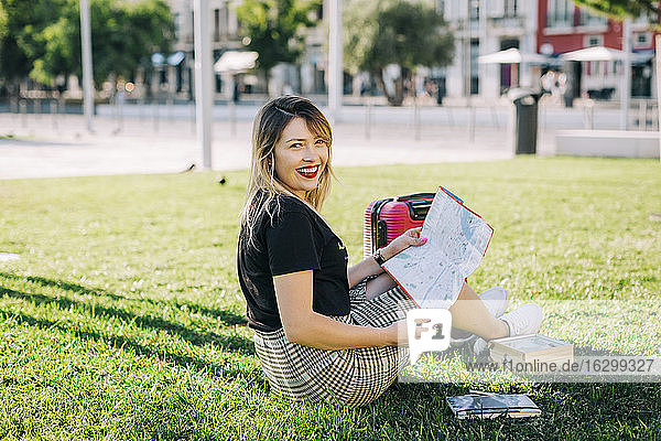Happy beautiful woman with map sitting on grassy land in park