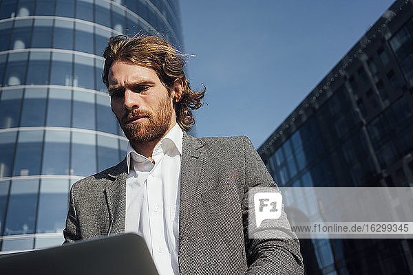 Bärtiger Geschäftsmann betrachtet Laptop im Finanzviertel an einem sonnigen Tag