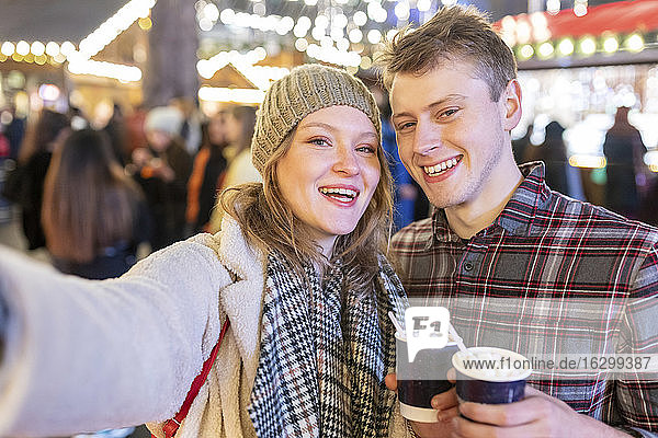 Glückliches Paar mit heißer Schokolade in der Hand auf einem beleuchteten Weihnachtsmarkt bei Nacht