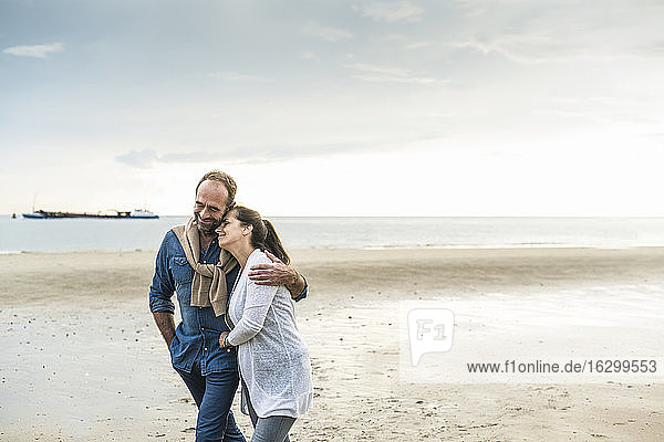 Mature man embracing woman while walking at beach against cloudy sky