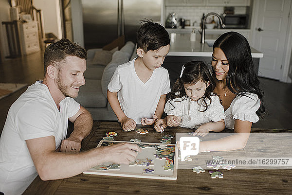 Lächelnde Eltern  die mit ihren Kindern am Tisch zu Hause ein Rätsel lösen