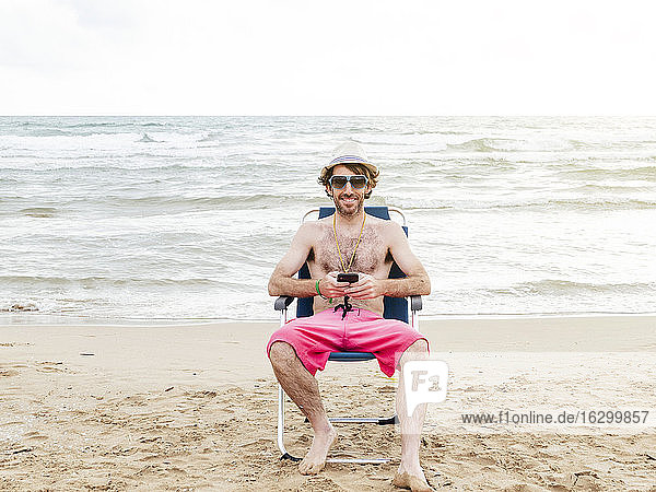 Smiling man using smartphone sitting in beach lounger on the beach