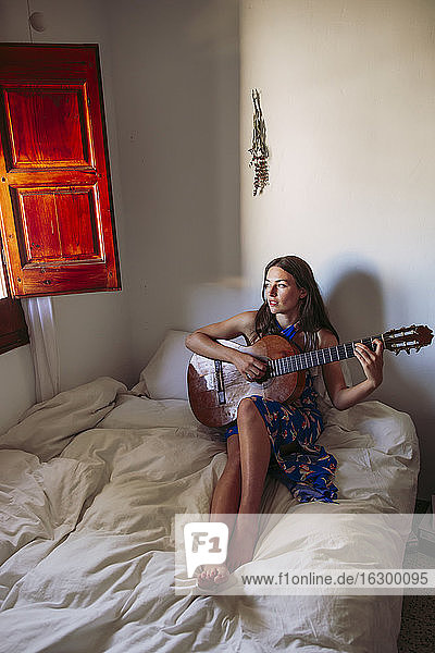 Junge Frau schaut weg  während sie zu Hause auf dem Bett Gitarre übt