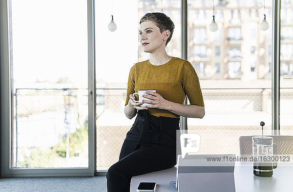Geschäftsfrau sitzt auf einem Schreibtisch im Büro und hält einen Kaffeebecher