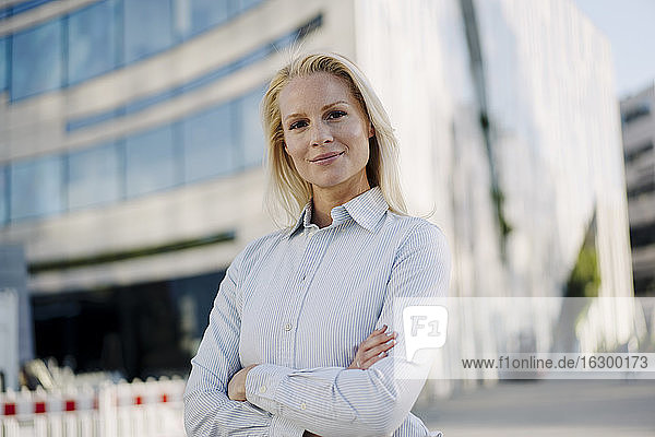 Beautiful blond female entrepreneur smiling while standing with arms crossed at financial district