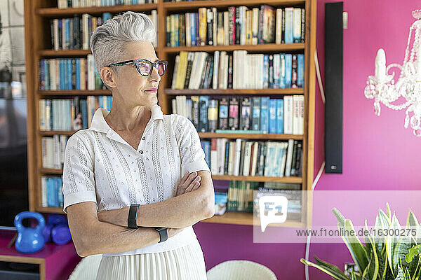 Ältere Frau mit verschränkten Armen  die zu Hause vor einem Bücherregal steht