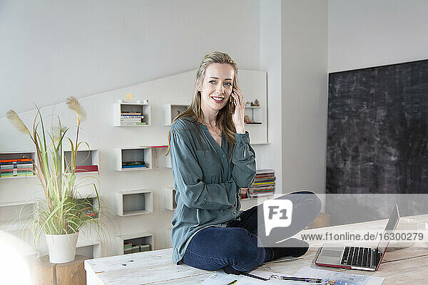 Woman sitting on desk in home office talking on the phone