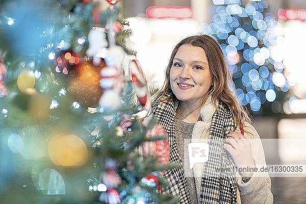Lächelnde schöne Frau  die an einem beleuchteten Weihnachtsbaum in einer nächtlichen Stadt steht