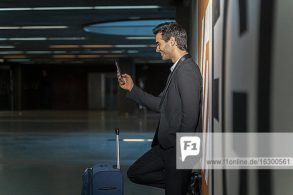 Lächelnder männlicher Unternehmer  der ein Mobiltelefon benutzt  während er mit einem Koffer an der Wand eines Bahnhofs steht