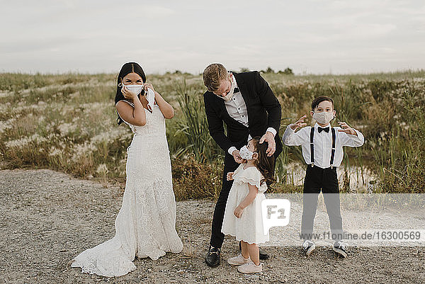 Eltern mit Kindern im Hochzeitskleid tragen einen Mundschutz  während sie während COVID-19 auf einem Feld stehen
