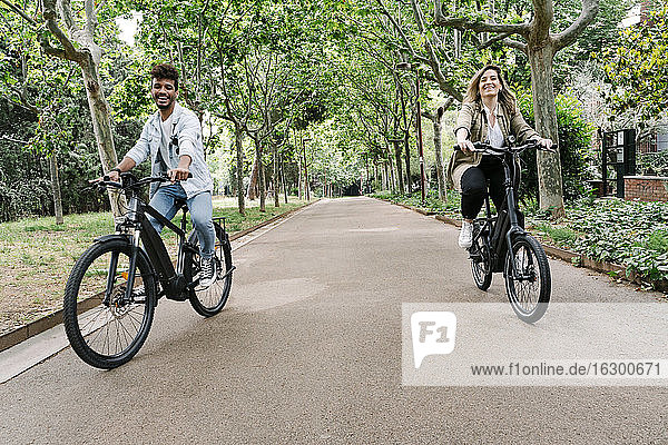 Happy young couple riding electric bicycles on road amidst trees
