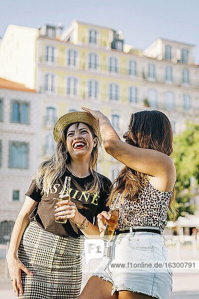 Young woman wearing hat to cheerful female friend while standing in city