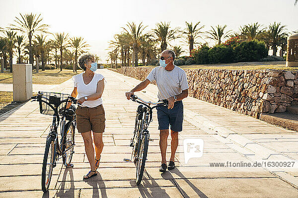Älterer Mann und Frau mit Gesichtsmaske unterhalten sich bei einem Spaziergang mit dem Fahrrad im Park
