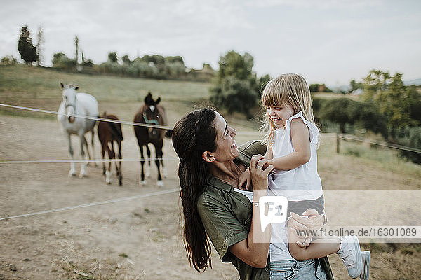 Lächelnde Mutter  die ihre fröhliche Tochter trägt  während sie auf dem Land gegen Pferde steht