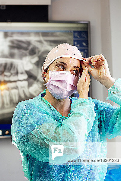 Dentist in protective suit wearing surgical cap while standing at clinic