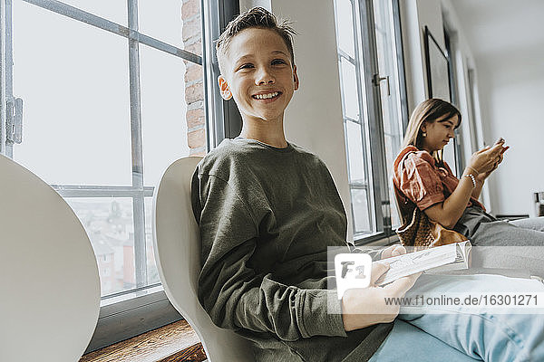 Lächelnder Junge mit Buch auf einem Stuhl sitzend  während ein Teenager-Mädchen im Wartezimmer ein Smartphone benutzt
