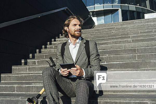 Geschäftsmann schaut weg  während er mit einem Tablet und einem Elektroroller auf einer Treppe im Finanzviertel sitzt