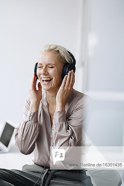 Fröhliche Geschäftsfrau hört Musik über Kopfhörer  während sie im Büro an der Wand sitzt