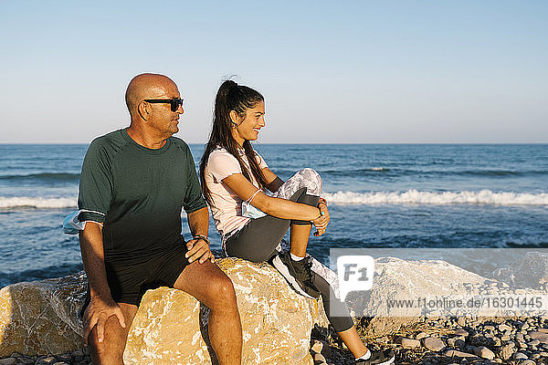 Senior man sitting by woman on rock against sea