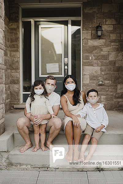 Familie mit Kindern trägt Schutzmaske  während sie auf den Stufen vor dem Haus sitzen