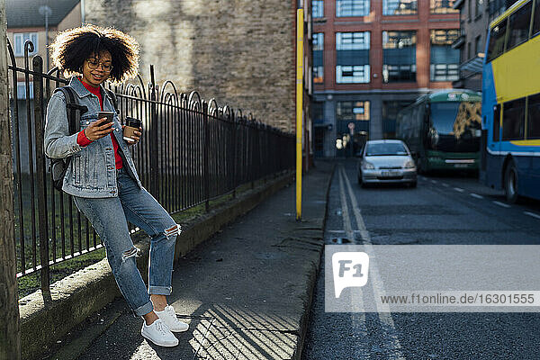 Afro young woman holding coffee cup using mobile phone while standing on street