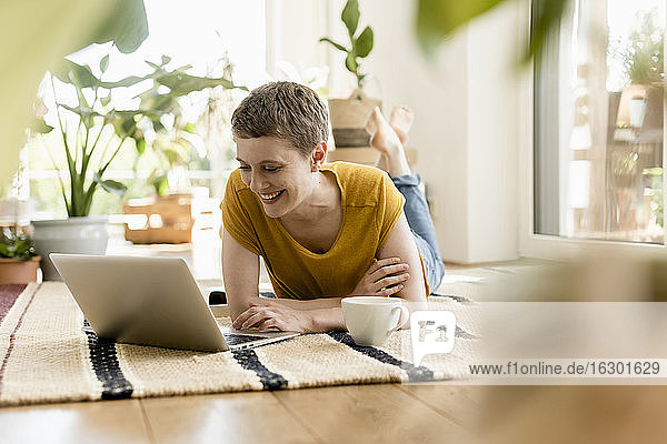 Lächelnde Frau im mittleren Erwachsenenalter  die einen Laptop benutzt  während sie zu Hause auf dem Teppich liegt