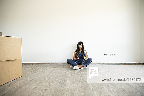 Schwangere Frau benutzt digitales Tablet  während sie auf dem Boden vor einer weißen Wand in einem neuen Haus sitzt