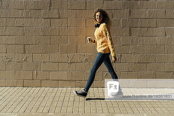 Junge Frau  die einen Einwegbecher hält  während sie an einem sonnigen Tag auf einem Fußweg an einer Mauer entlang läuft