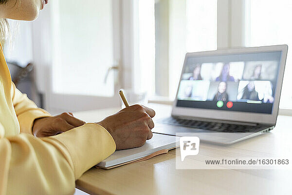 Abgeschnittene Hände einer Unternehmerin  die in ein Tagebuch schreibt  während sie mit Kollegen per Videokonferenz zu Hause spricht